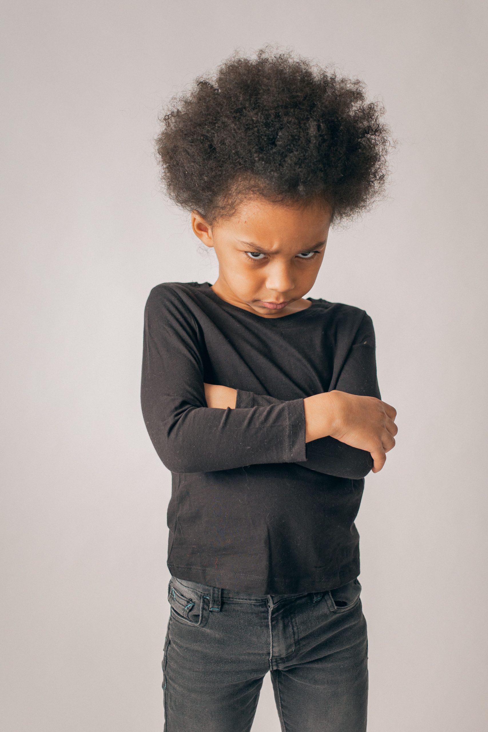 Unhappy African American girl in black clothes with Afro hairstyle standing on white background with crossed arms in light room